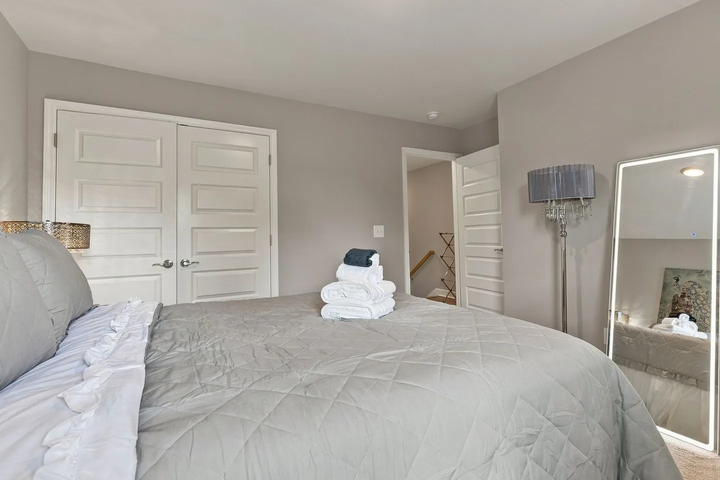 Bedroom with a bed made with a grey quilt and folded towels, featuring closet doors and a full-length lighted mirror.