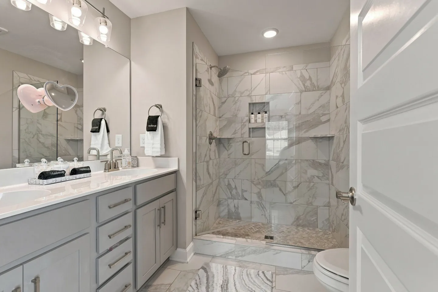 Bathroom featuring a gray double vanity, large mirror, and a walk-in glass shower with marble-style tile walls.