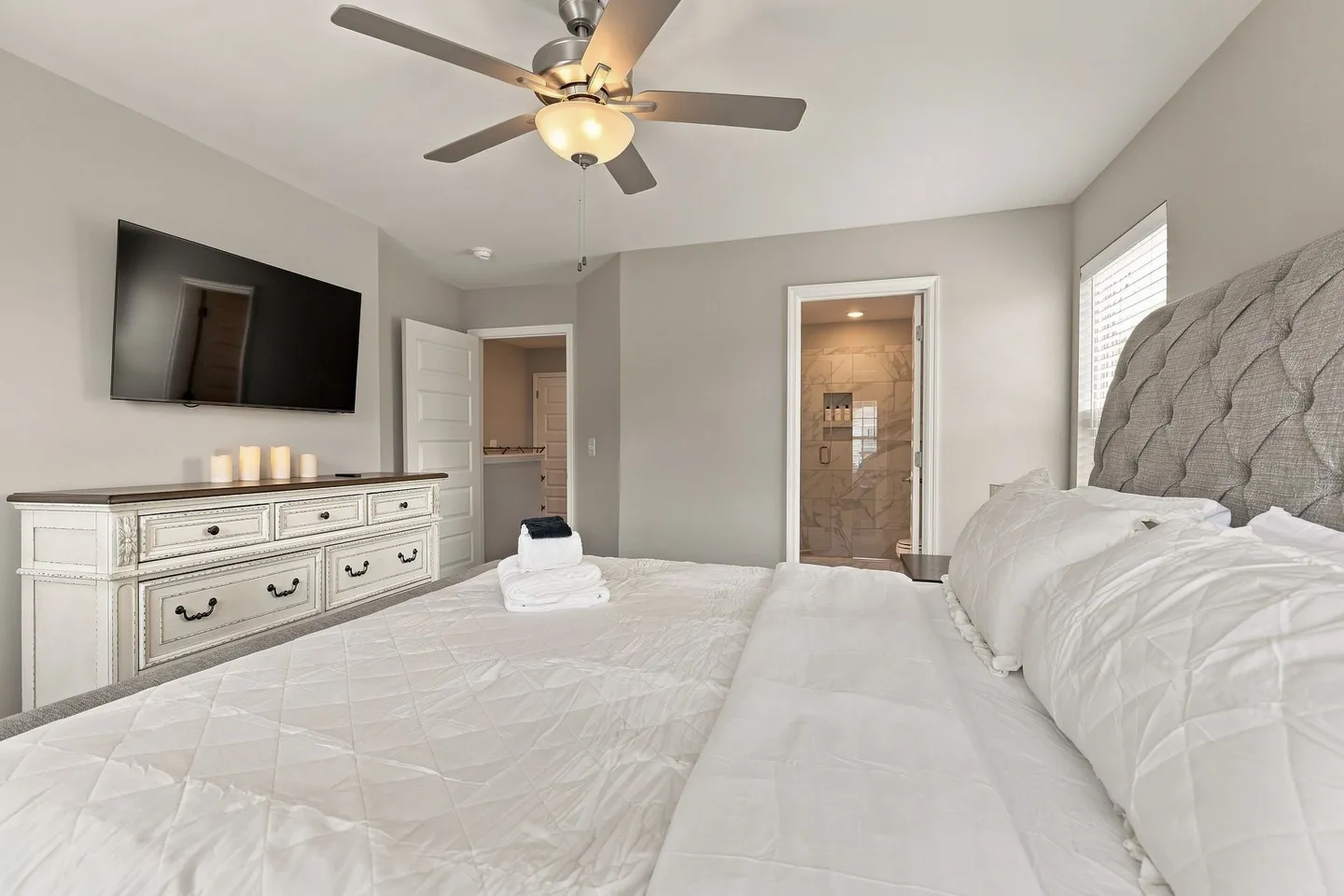 Bedroom featuring a bed with a tufted headboard, white dresser beneath a wall-mounted TV, and an open doorway to an ensuite bathroom.