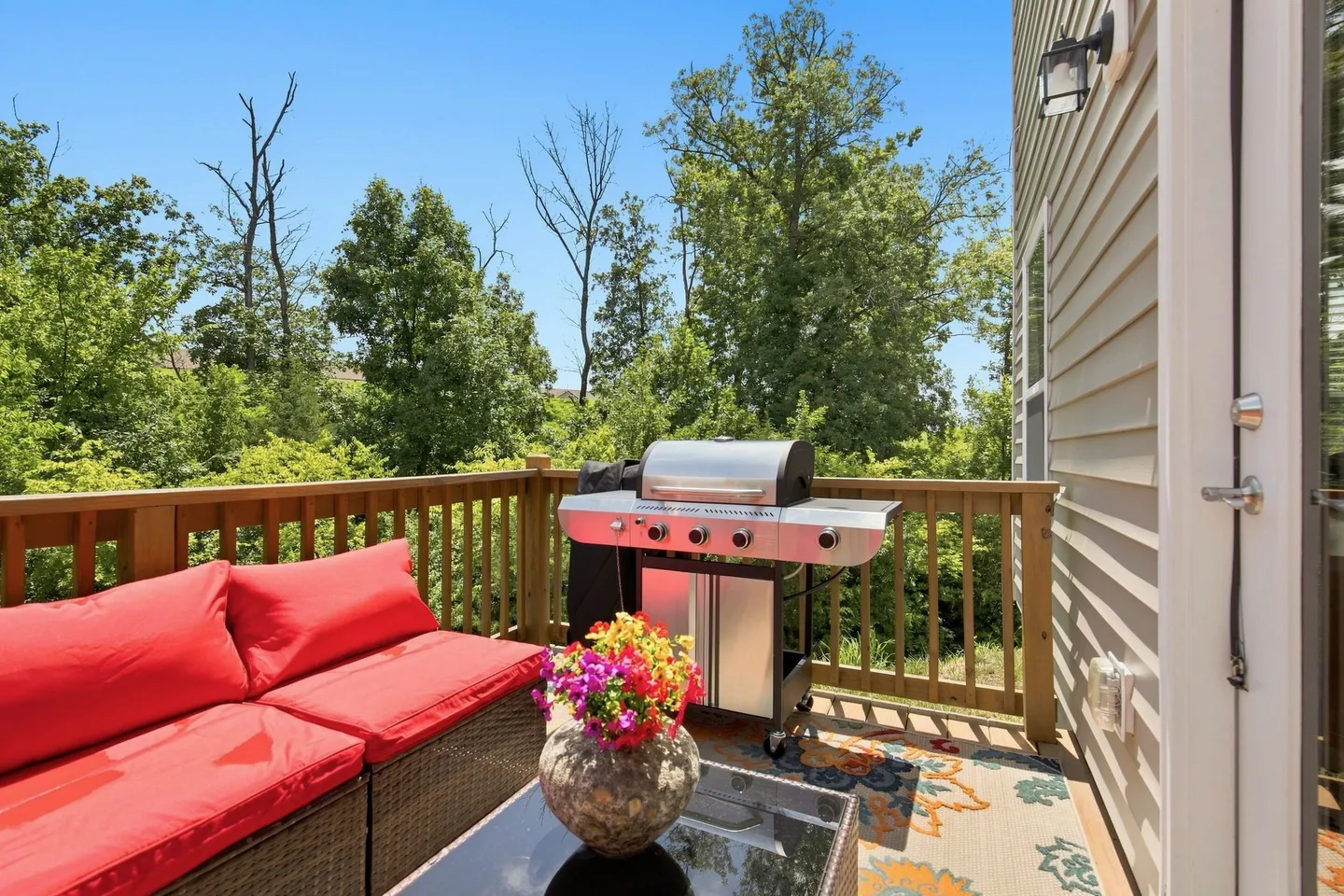 Outdoor wooden deck featuring a wicker sofa with bright red cushions, a glass-top coffee table with flowers, and a stainless steel barbecue grill against a wooden railing overlooking green trees.
