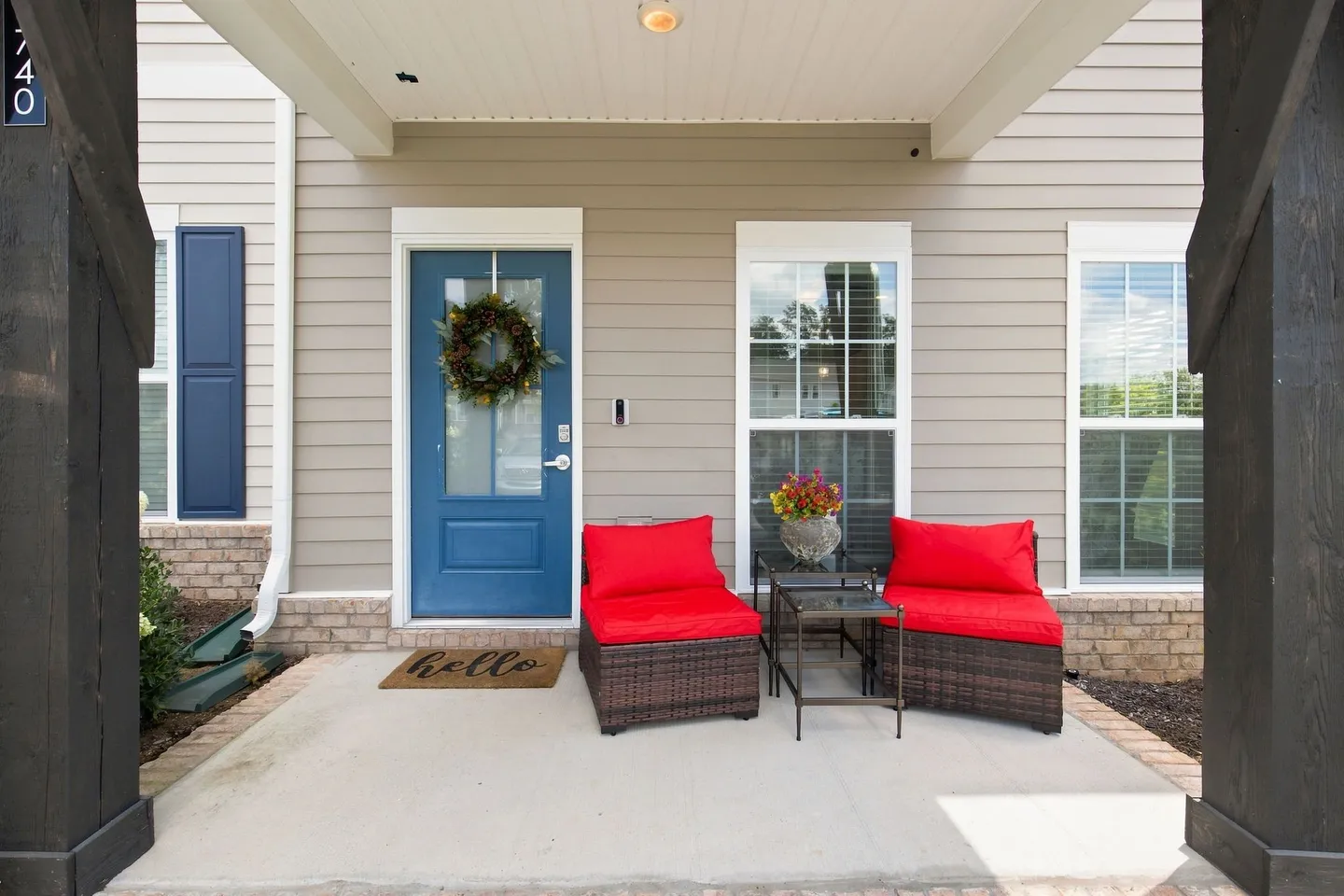 Front porch of a house featuring a blue front door with a wreath, a welcome mat, and two wicker chairs with bright red cushions.