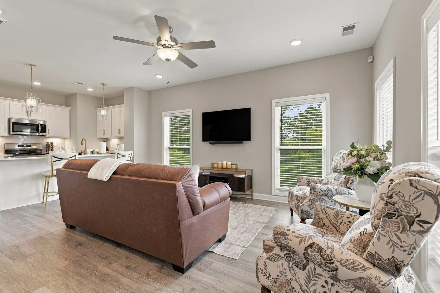 Open living area with brown leather sofa, patterned armchairs, wall-mounted TV, and kitchen in the background.