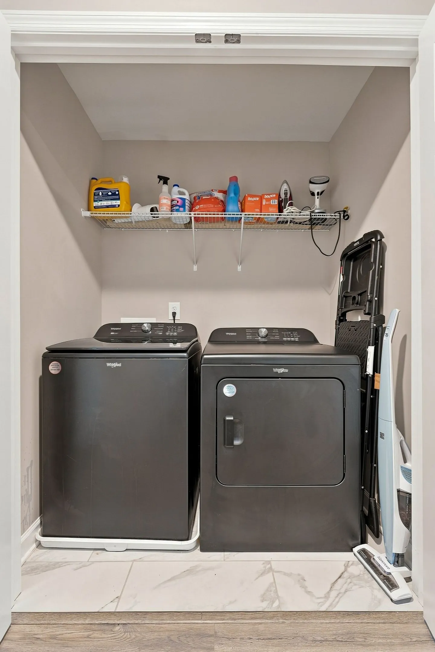 A laundry closet featuring a dark grey Whirlpool washing machine and dryer beneath a wire shelf stocked with cleaning supplies.
