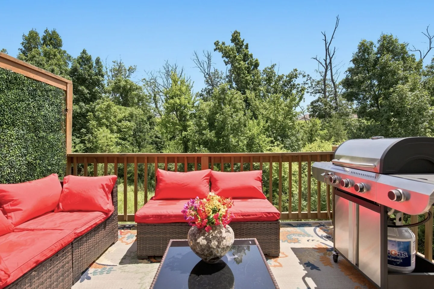Outdoor wooden deck featuring wicker seating with red cushions, a gas barbecue grill, and a green tree backdrop under a clear blue sky.