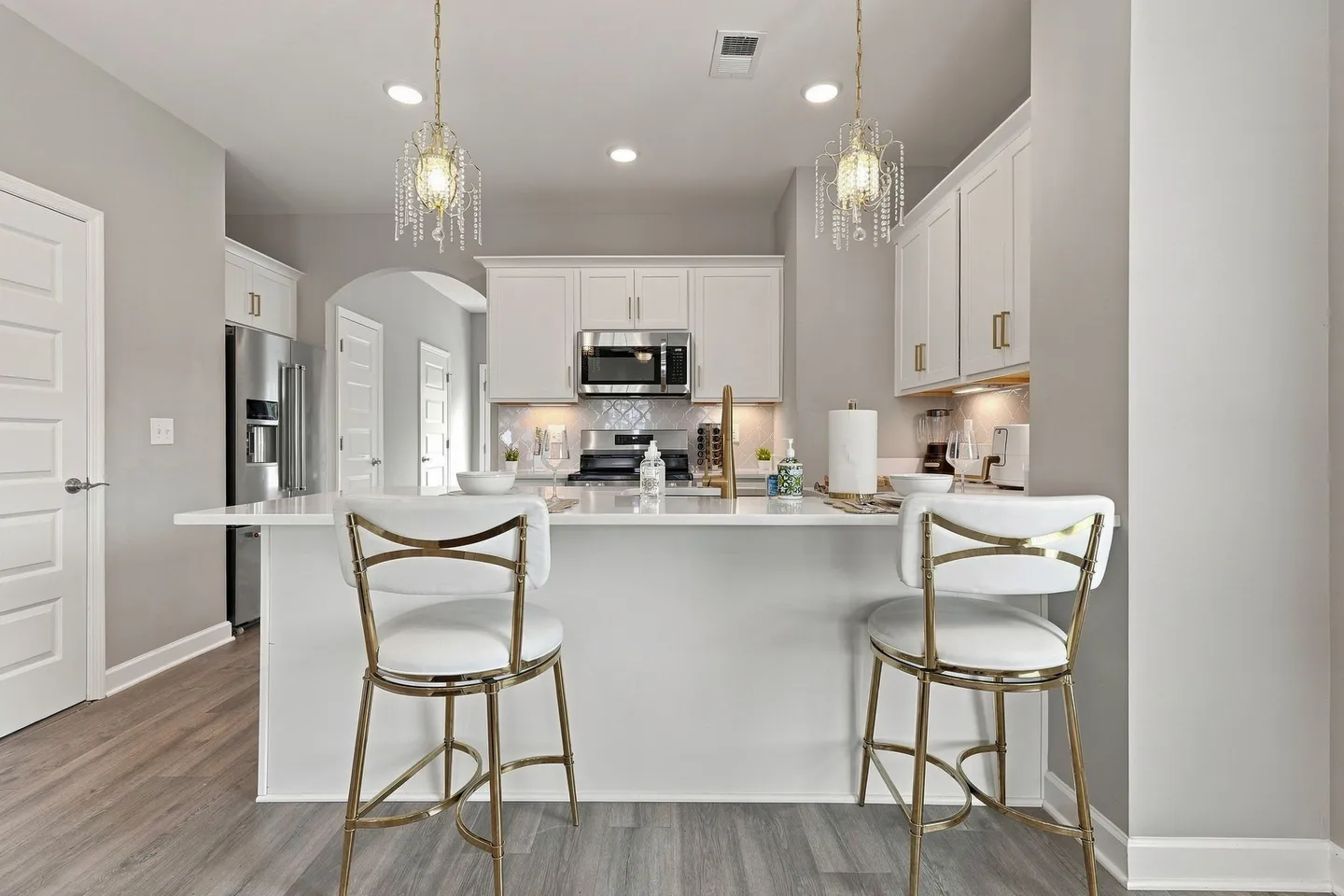 Kitchen island with two white and gold barstools beneath crystal pendant lights, leading into a white-cabinet kitchen with stainless steel appliances.