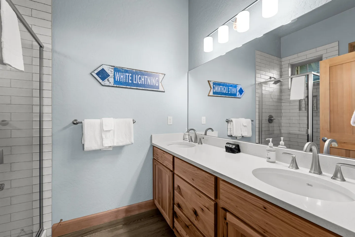 Bathroom with a double-sink wood vanity, large mirror with vanity lighting, and a glass-enclosed subway tile shower.