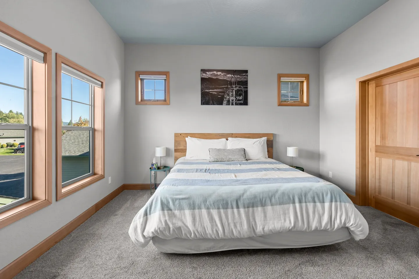 Bedroom with carpeted floor, light-colored walls, soft blue ceiling, a bed with a wooden headboard, two small high windows, and two larger windows.