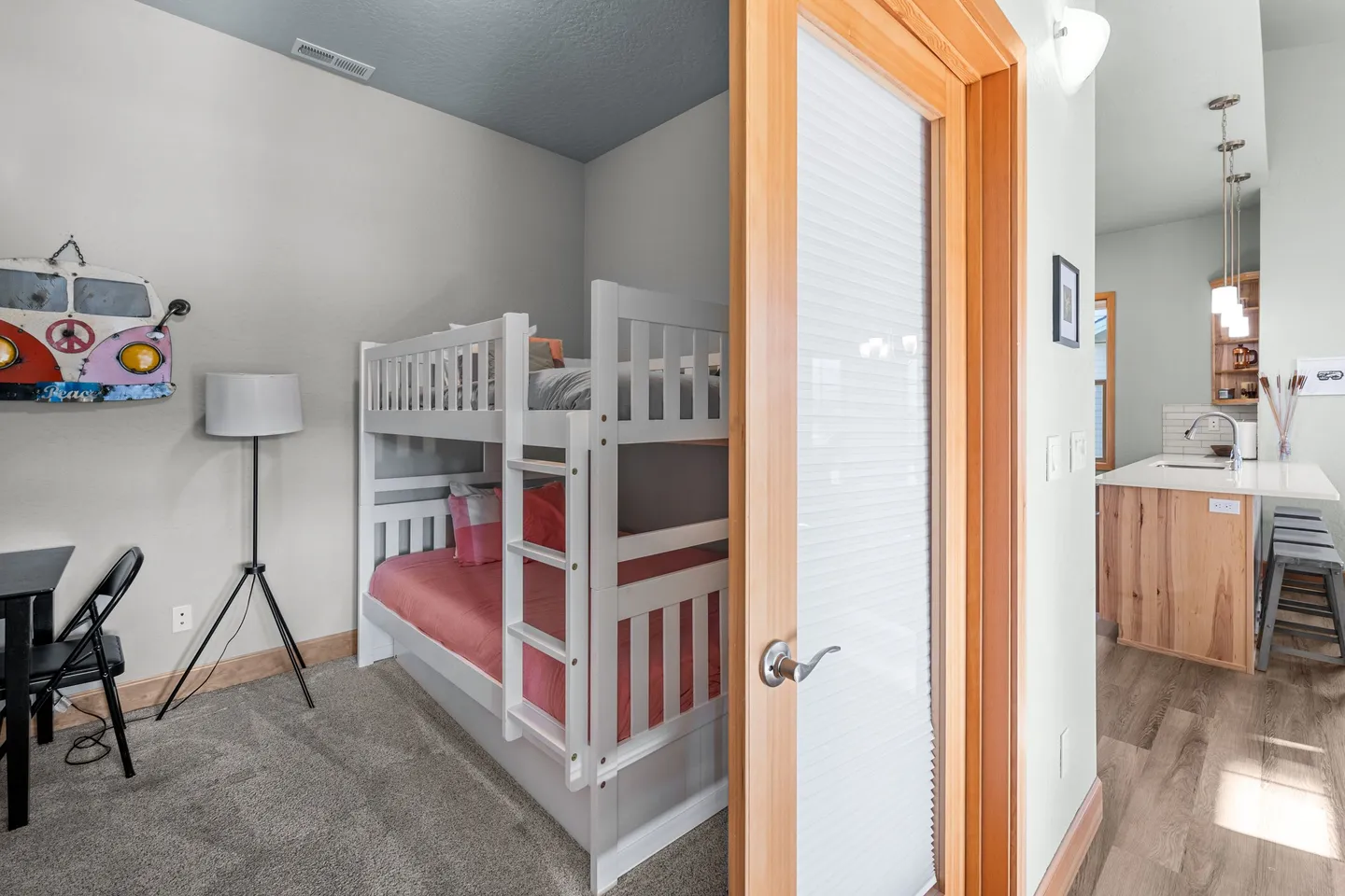 View through an open doorway of a bedroom with white wooden bunk beds and a small desk, with a kitchen visible in the adjacent hallway.