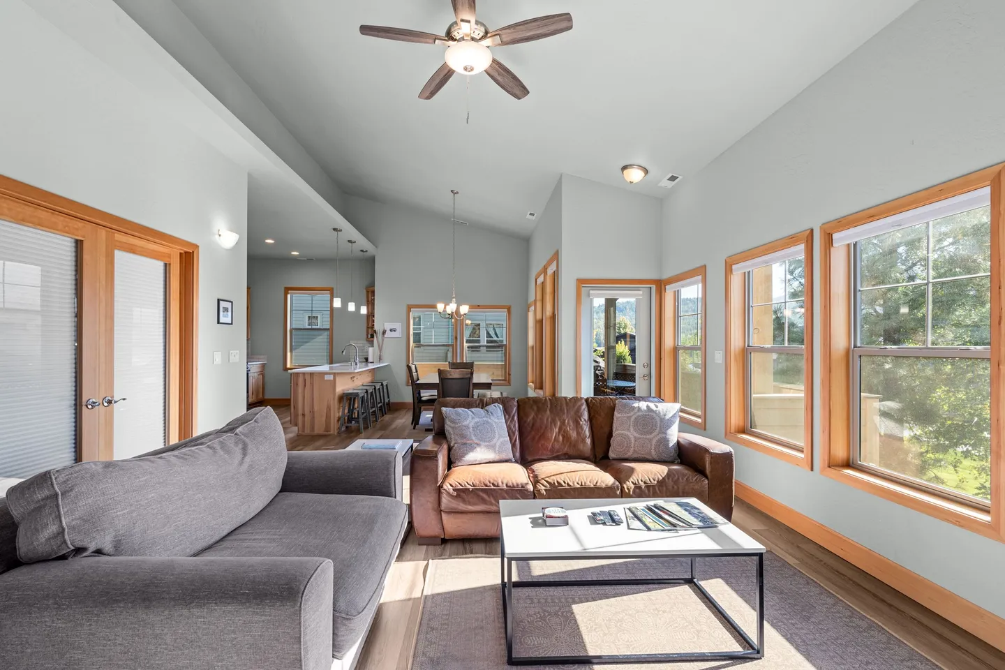Open-concept living room with gray and brown sofas, coffee table, vaulted ceiling, and view into the kitchen and dining area.