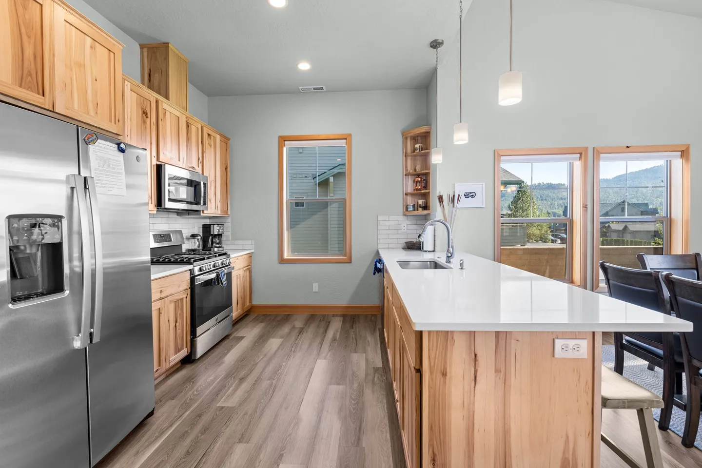 Bright kitchen featuring light wood cabinetry, stainless steel appliances, a white counter kitchen island with pendant lights, and wood-look flooring.
