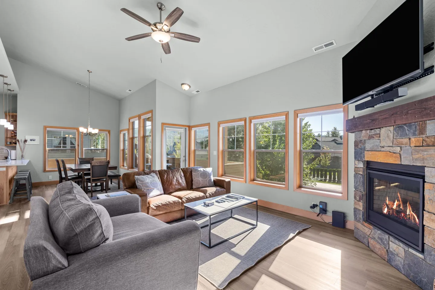 Living room with vaulted ceiling, leather sofa, armchair, fireplace with stone surround, wall-mounted TV, and large windows.