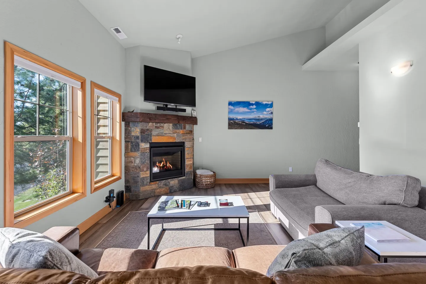 Living room with vaulted ceiling, corner stone fireplace with mounted TV, windows, and sofa seating around a coffee table.