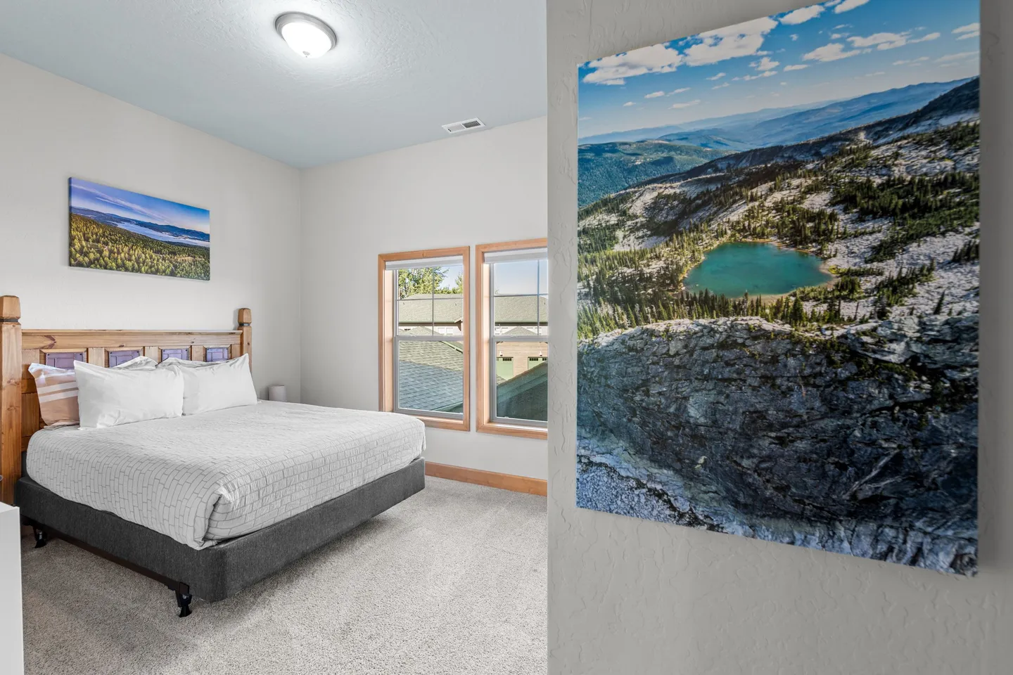 Bedroom with a wooden headboard, made bed, carpet flooring, two windows, and framed nature art on the walls.