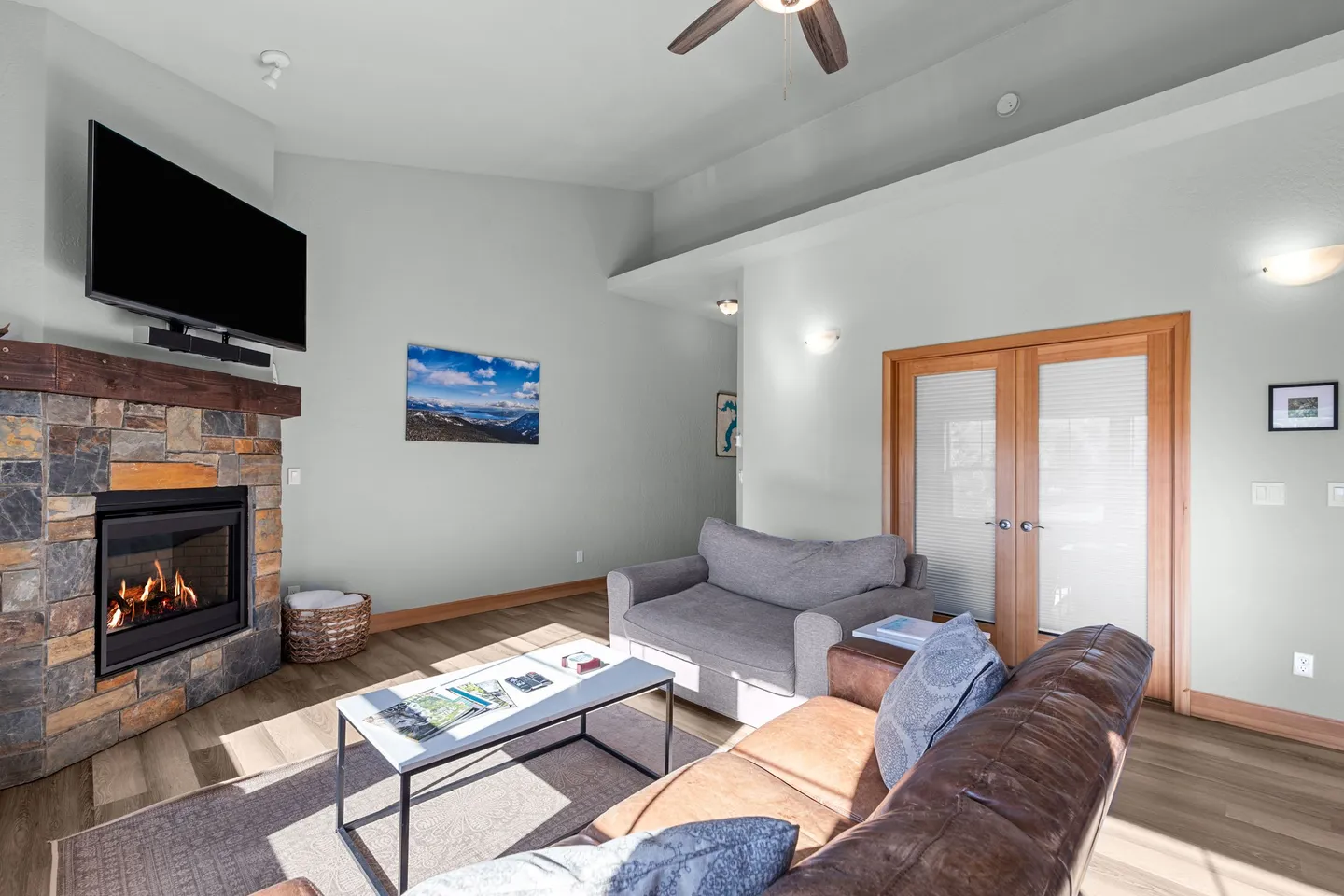 Living room with vaulted ceiling, stone-surround fireplace, wall-mounted TV, leather couch, and double doors.