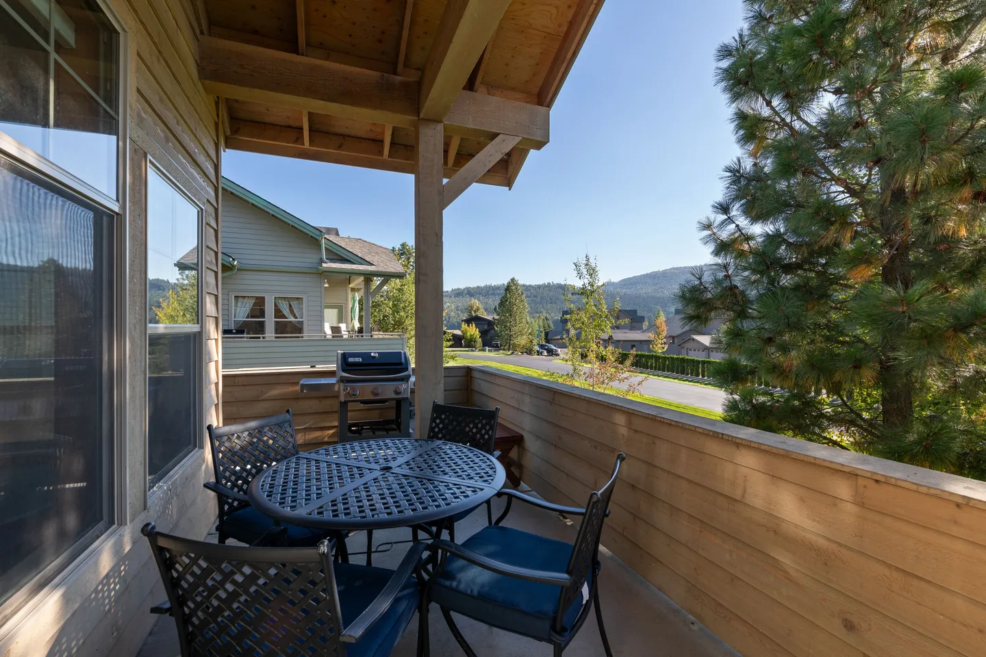 Covered outdoor balcony with a round patio table, chairs with blue cushions, and a barbecue grill overlooking a street and trees.