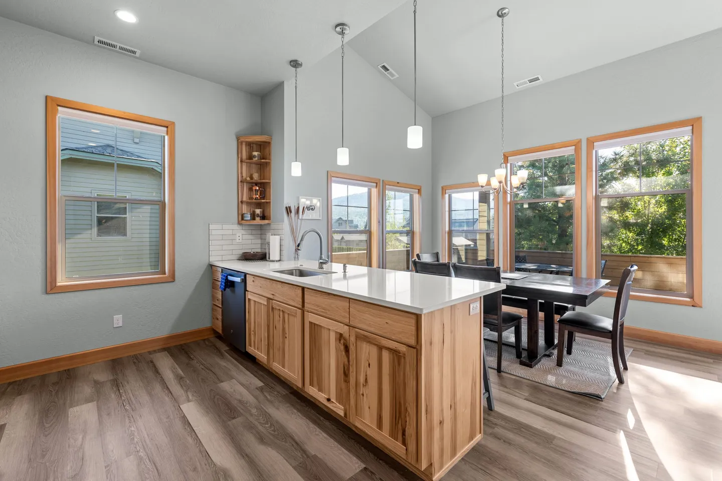 Open-concept kitchen and dining area featuring a light wood island, white countertops, vaulted ceilings, and a dining table with chairs next to large windows.