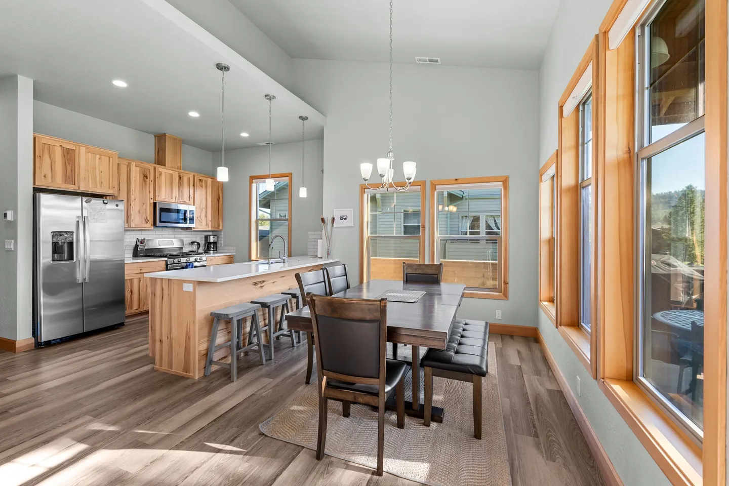 Open-concept dining area and kitchen with wood cabinetry, stainless steel appliances, a dining table with bench and chairs, and wood-framed windows.