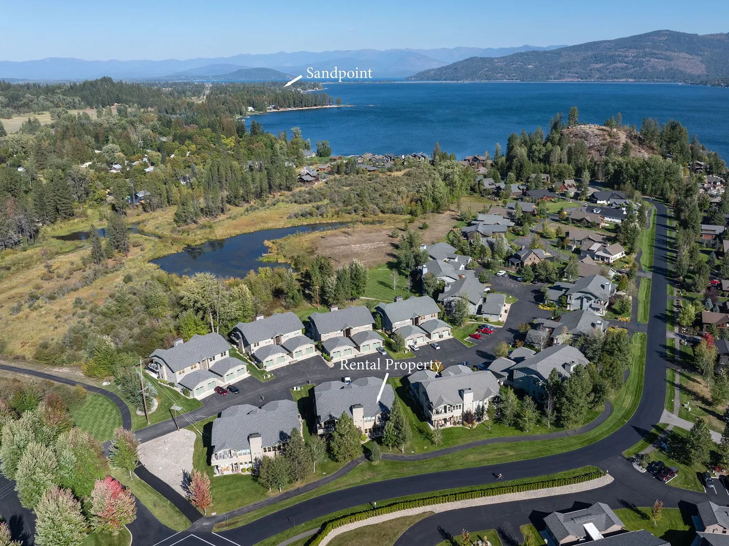 Aerial view of a residential community of multi-story townhomes with a nearby pond, large lake, and distant mountains under a blue sky.