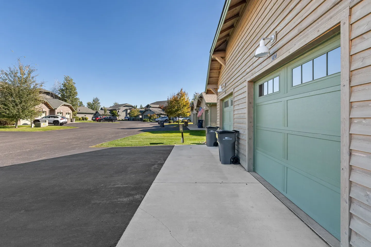 Exterior garage side of a wood-sided townhome showing green garage doors, trash bins, and a paved neighborhood street under a clear blue sky.