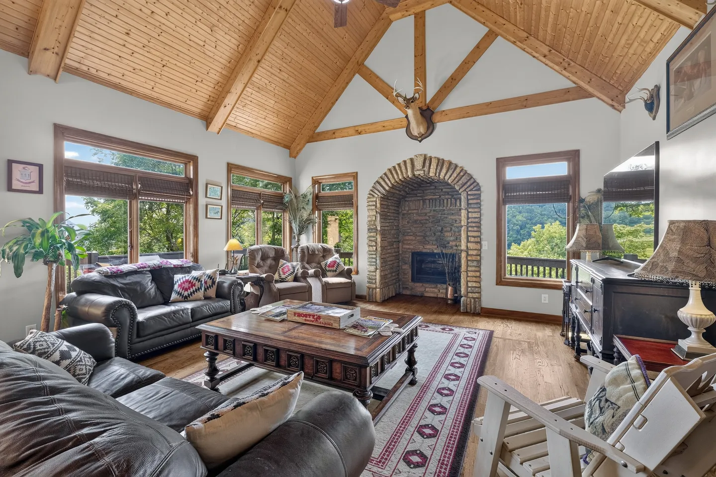 Living room with high wood-paneled vaulted ceiling, stone fireplace, leather sofas, coffee table, and windows showing outdoor views.