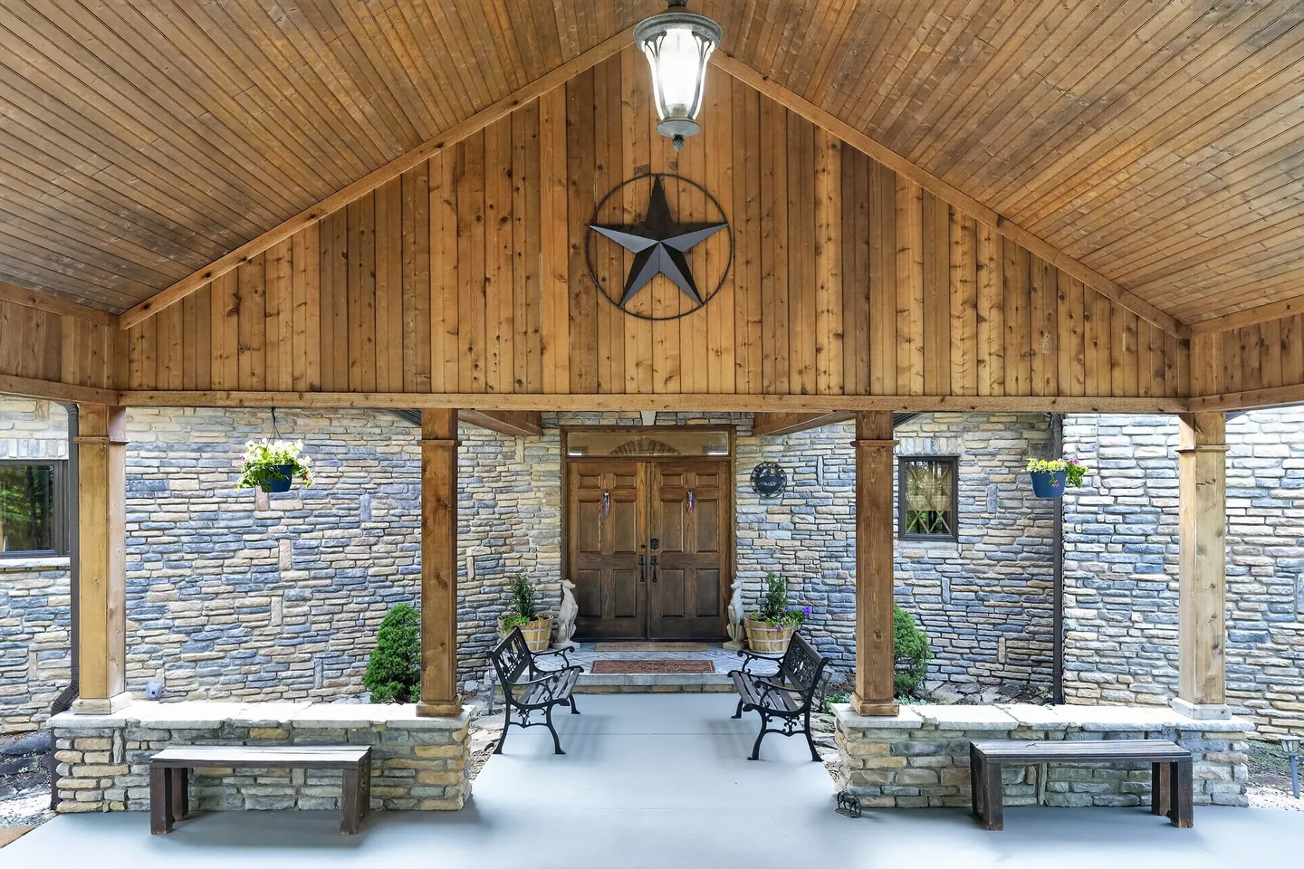 A covered stone front porch with wood-paneled gable ceiling, metal star decor, hanging pendant light, benches, and double wooden front doors.