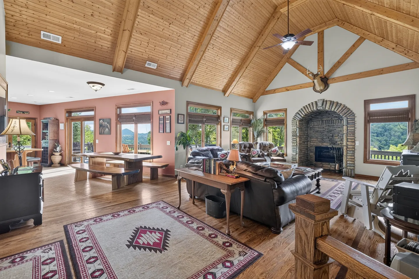 Open-concept living and dining area featuring vaulted wood ceilings, a stone fireplace, leather seating, and mountain views through wide windows and glass doors.