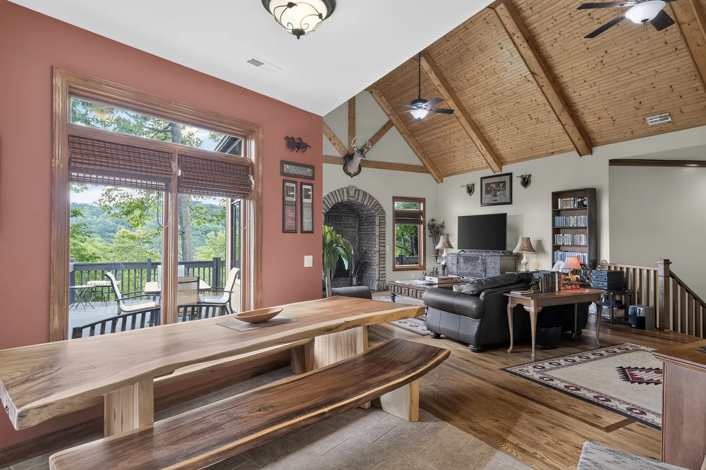 Open living and dining area featuring a live-edge wooden table and bench, vaulted wood-paneled ceiling with exposed beams, leather sofa, and sliding glass door leading to an outdoor deck.