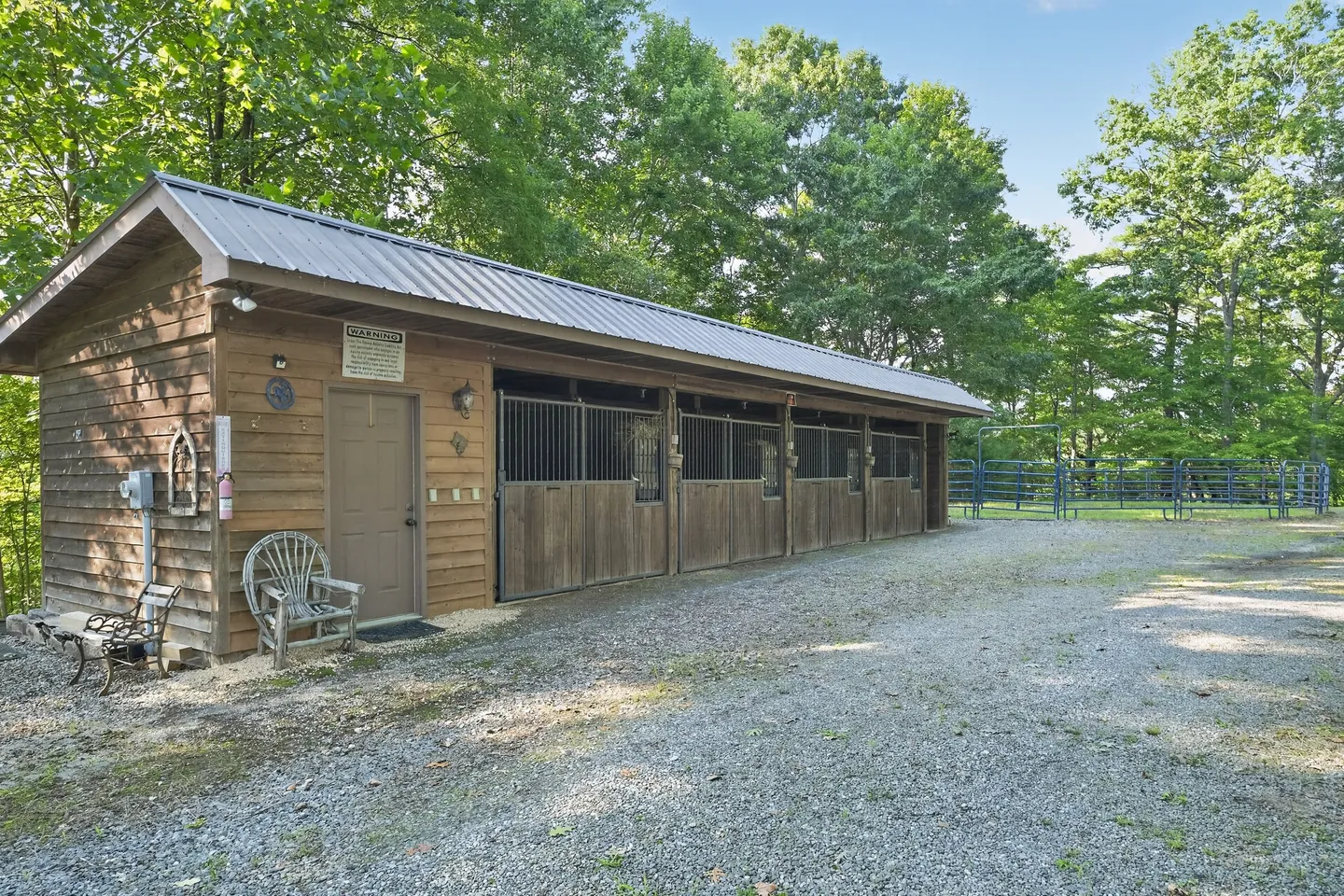 Exterior view of a wooden horse barn with metal roof and stalls alongside a gravel driveway and round pen.
