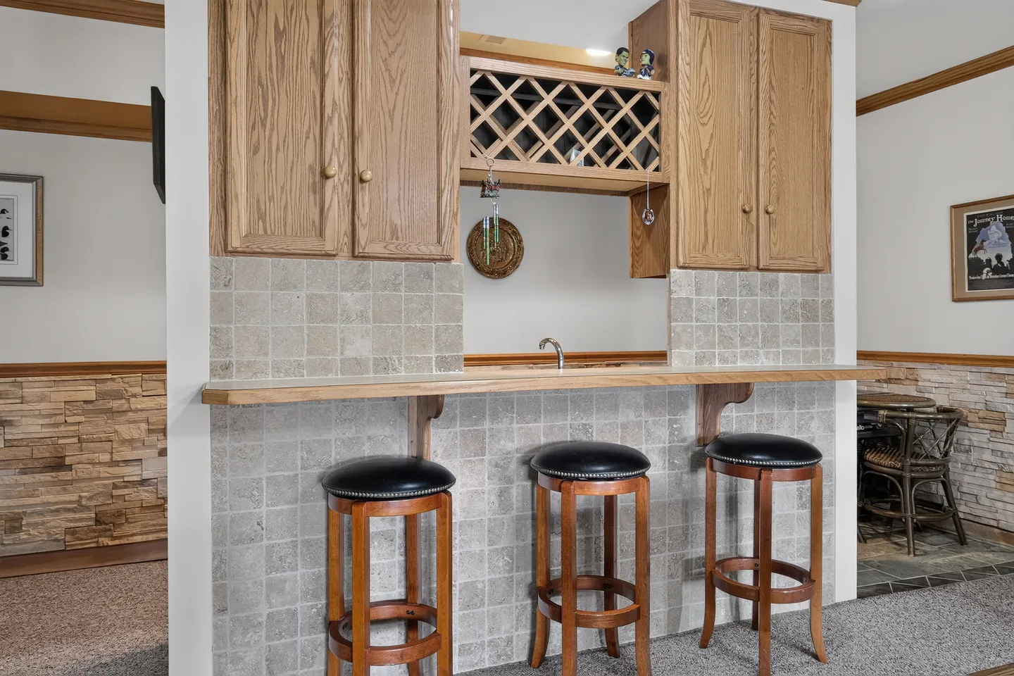 A built-in bar counter tiled in light grey stone with three barstools, wood cabinetry, and a wine rack.