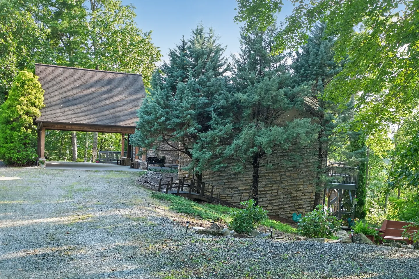 Exterior of a stone house with an attached carport, gravel driveway, and tall evergreen trees.