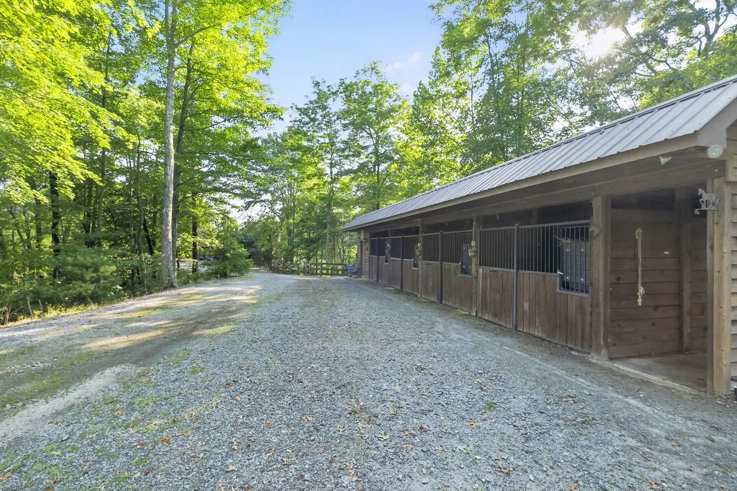 A long wooden horse barn with open-front stalls alongside a wide gravel drive surrounded by tall green trees.