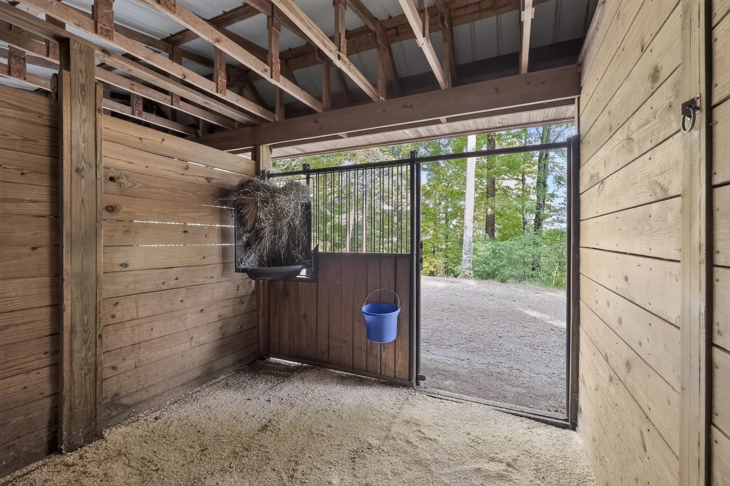 Interior view of a horse stall with wood-paneled walls, an open metal-barred gate, a hay feeder, and wood shavings on the floor looking out toward a gravel driveway.