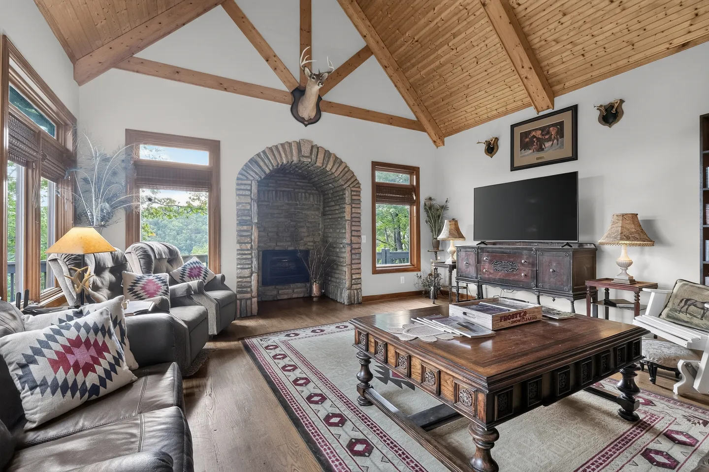 Living room with vaulted wood-paneled ceiling, exposed beams, stone fireplace nook, leather seating, and a large carved wooden coffee table.