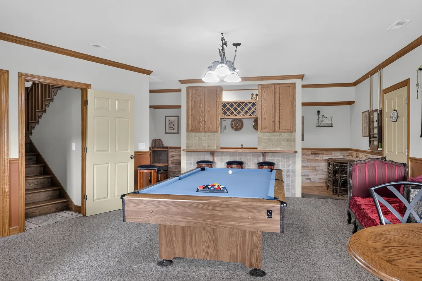 Game room featuring a light blue felt pool table, carpeted flooring, and a wet bar with wooden cabinetry and barstools in the background.
