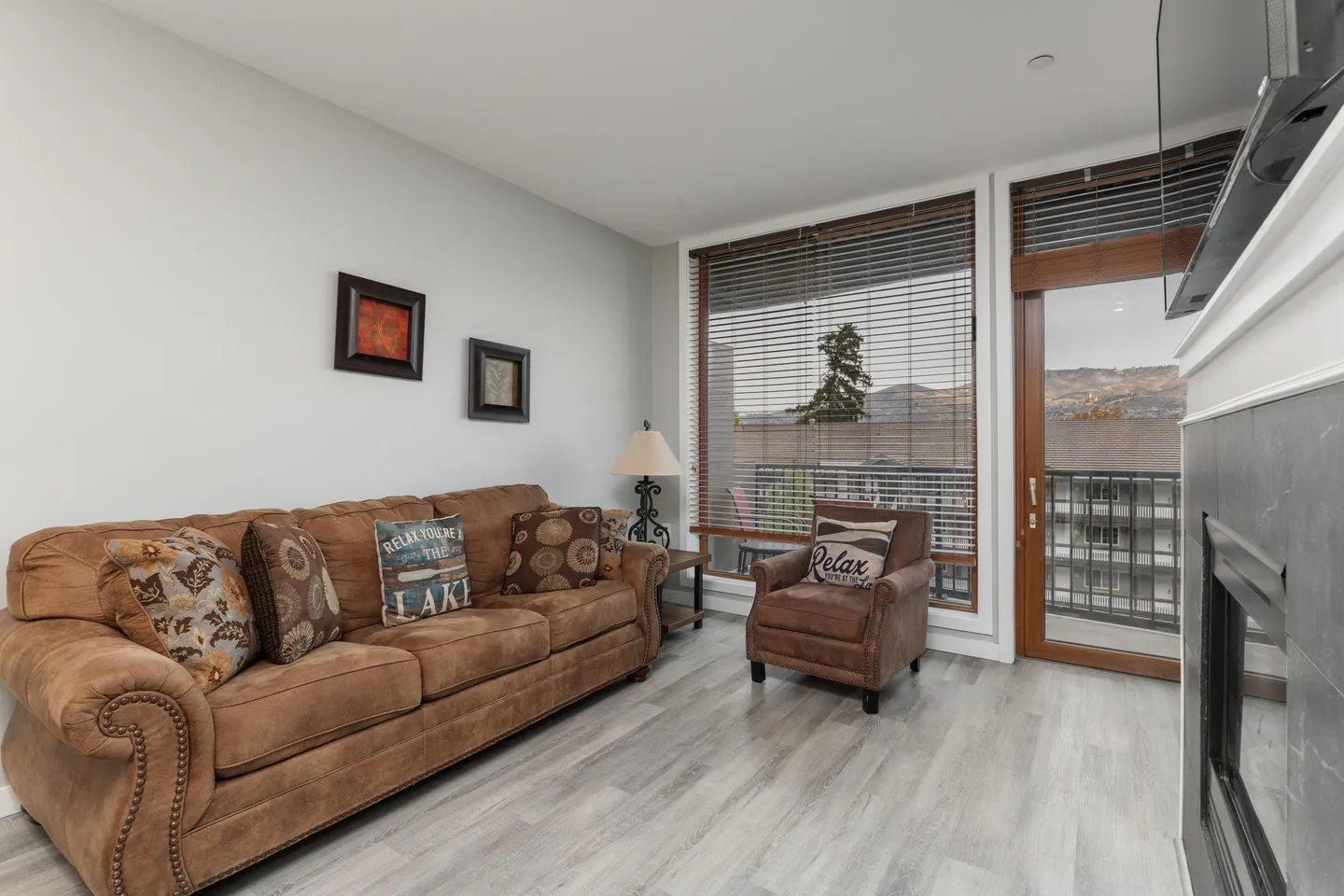 Living room with brown sofa, armchair, fireplace edge, and glass door leading to a balcony.
