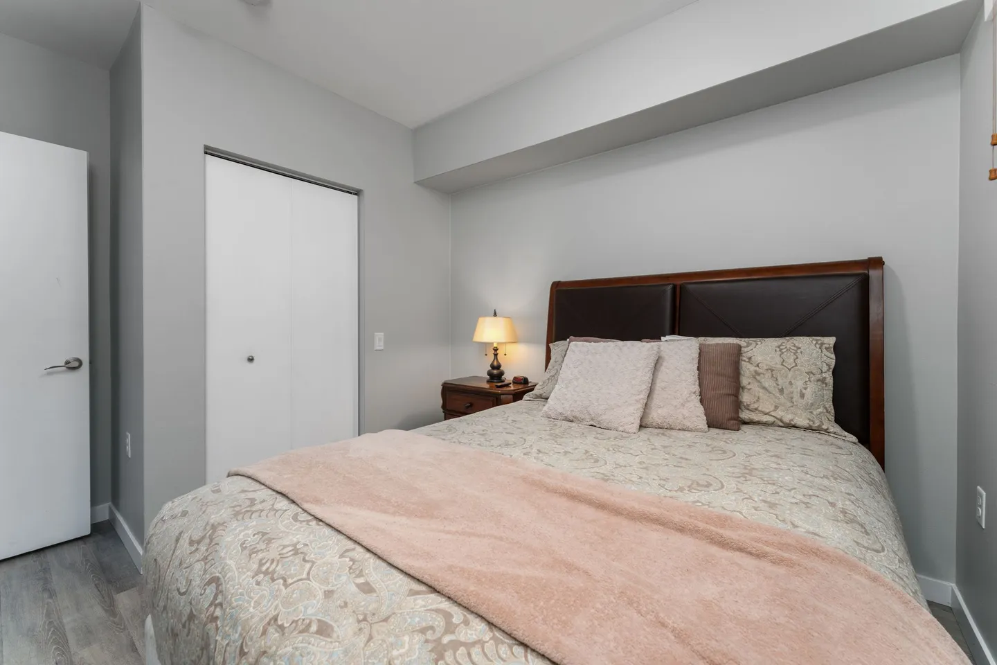 Bedroom with a made bed, dark wood headboard, bedside table with a lighted lamp, and white closet door.