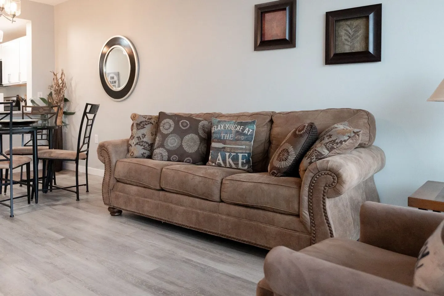 Living room featuring a brown upholstered sofa with throw pillows, wood-look flooring, and wall art, with a dining table visible nearby.