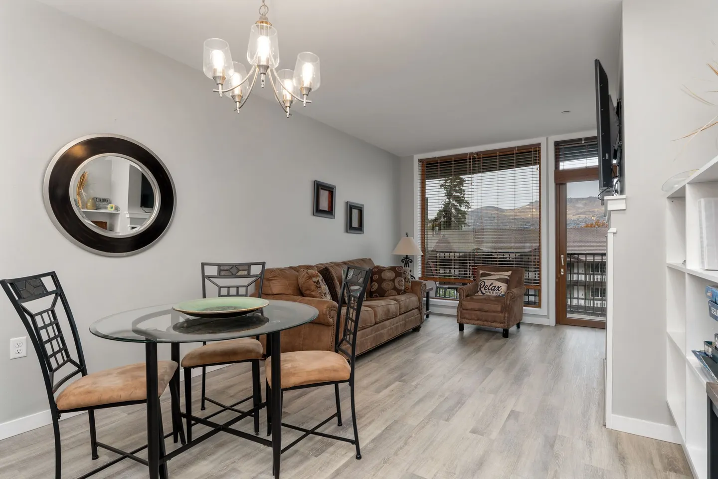 Open living and dining area featuring a round glass table with three chairs, a brown sofa, and a glass door leading to a balcony.