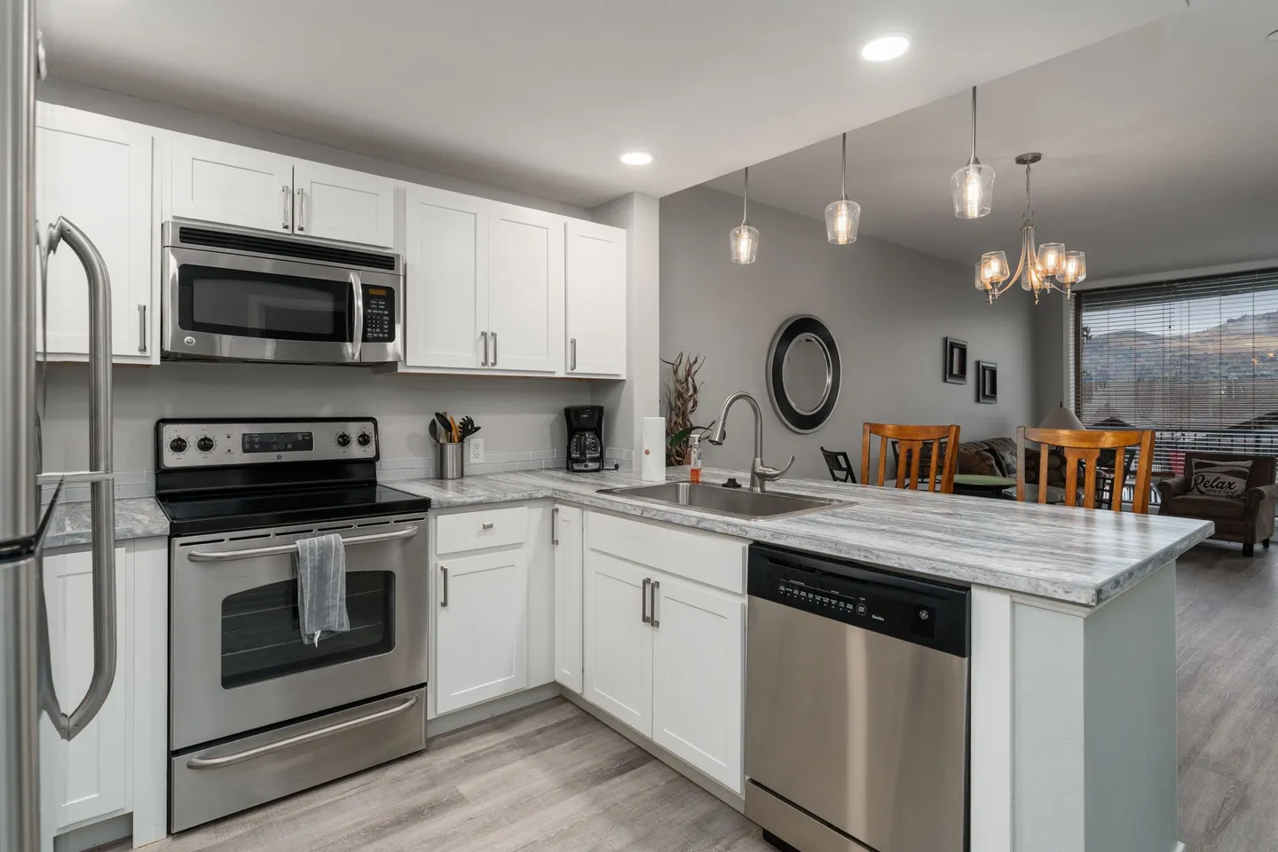 Kitchen with white shaker cabinets, marbled countertops, and stainless steel appliances opening into a living and dining area.
