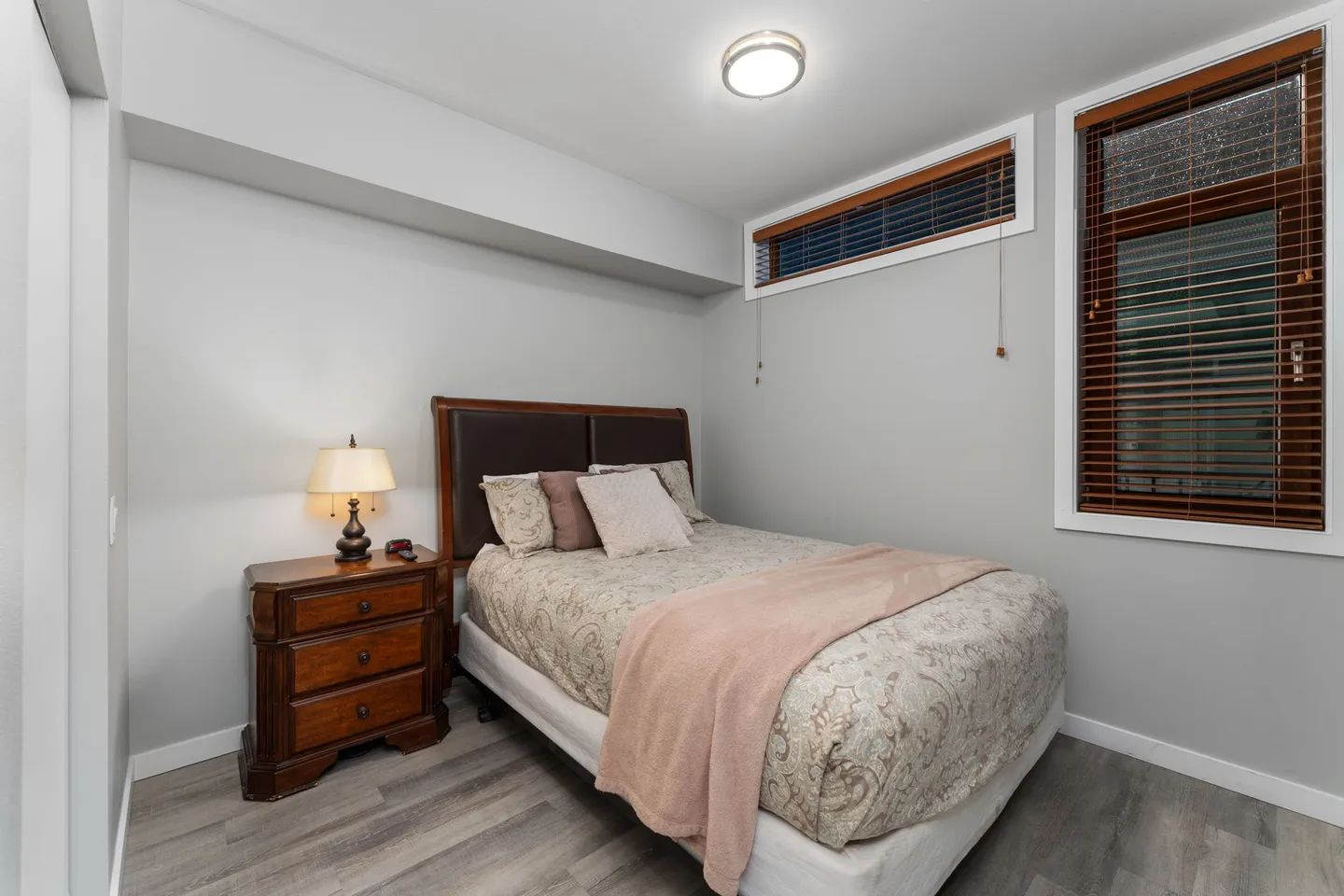 Bedroom with a made bed, wood headboard, nightstand with a lamp, and high windows with blinds.