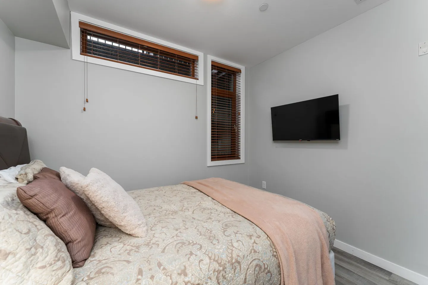 Bedroom with a made bed, two high-set windows with wooden blinds, and a wall-mounted flat-screen TV.