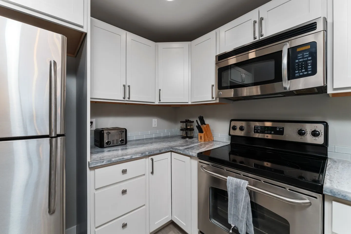Kitchen corner showing white shaker cabinets, marbled countertops, stainless steel refrigerator, microwave, and electric range.