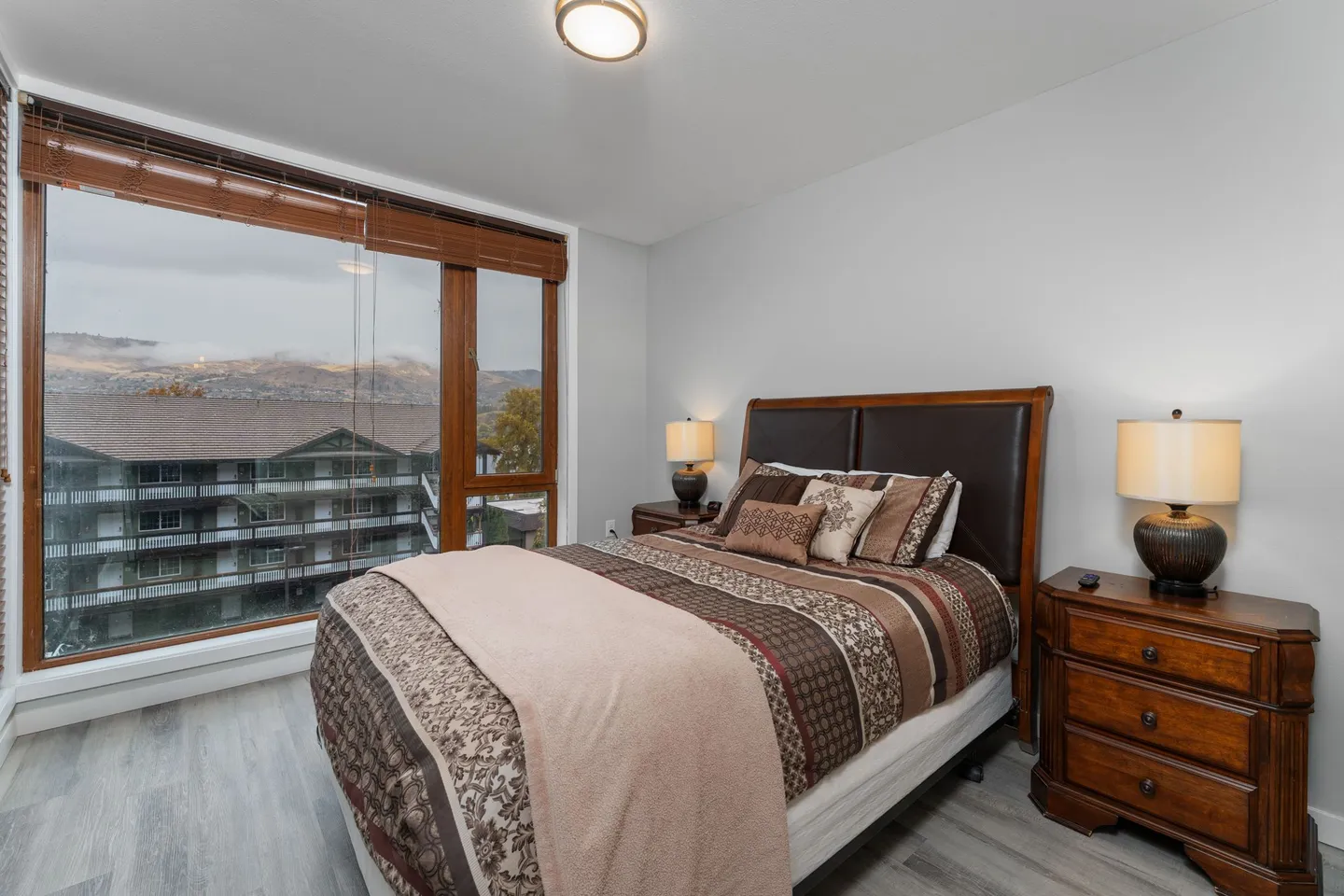 Bedroom with a wooden frame bed, matching nightstands with lamps, and large corner windows looking out at nearby buildings and hills.