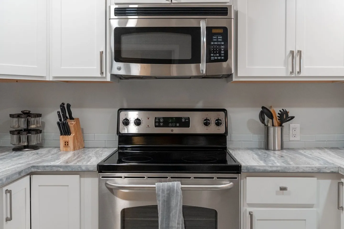 Close-up of a kitchen cooking area with a stainless steel range, built-in microwave, white shaker cabinets, and marble-look countertops.
