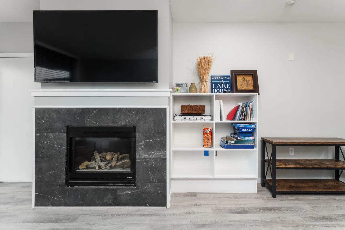 A living room fireplace with dark tile surround, a wall-mounted TV above the mantle, and a white bookcase filled with games.