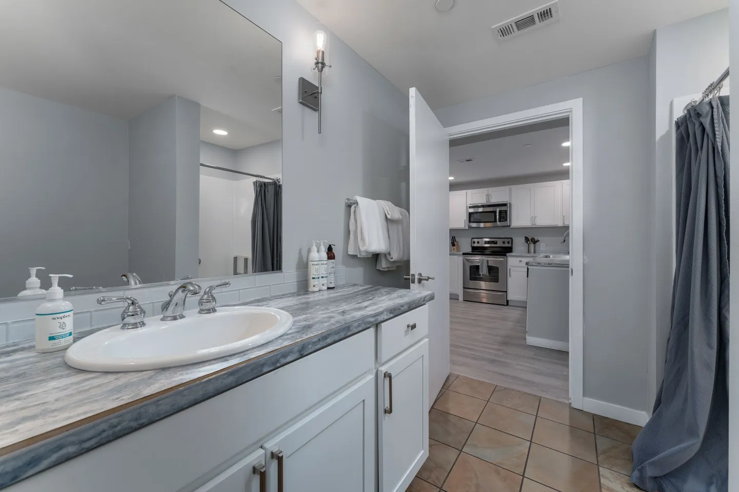 Bathroom with white vanity, swirl-patterned countertop, tiled floor, and an open doorway showing the kitchen.