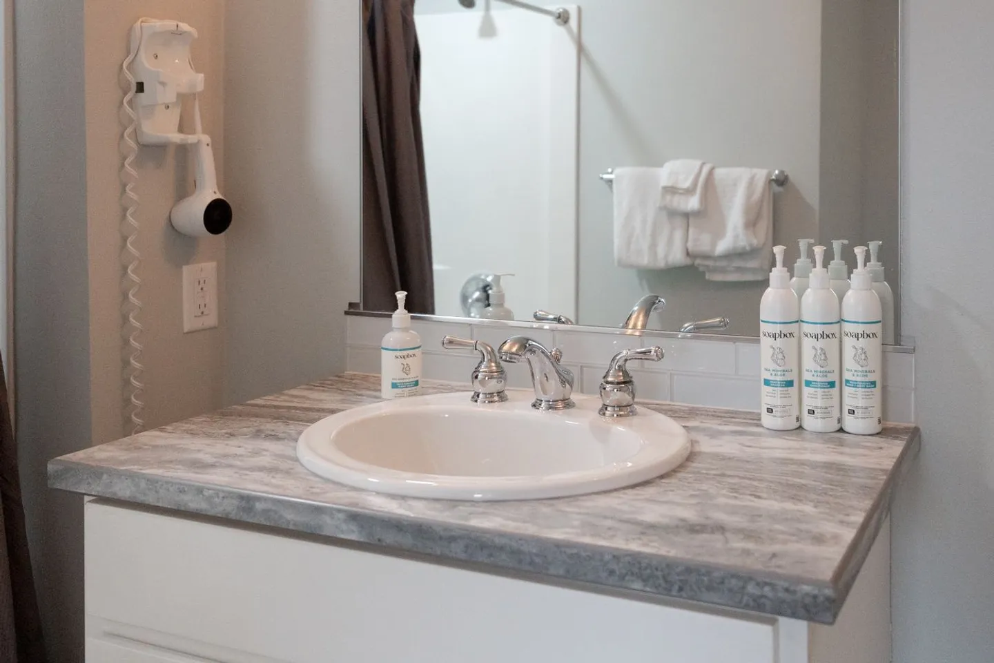Bathroom vanity with an oval sink, marbled countertop, wall-mounted hair dryer, mirror, and toiletries.