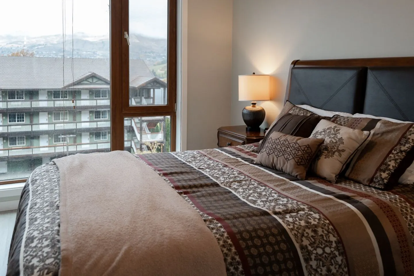 Bedroom with a patterned comforter and pillows on the bed next to a nightstand with a lamp and a tall window overlooking nearby buildings and hills.