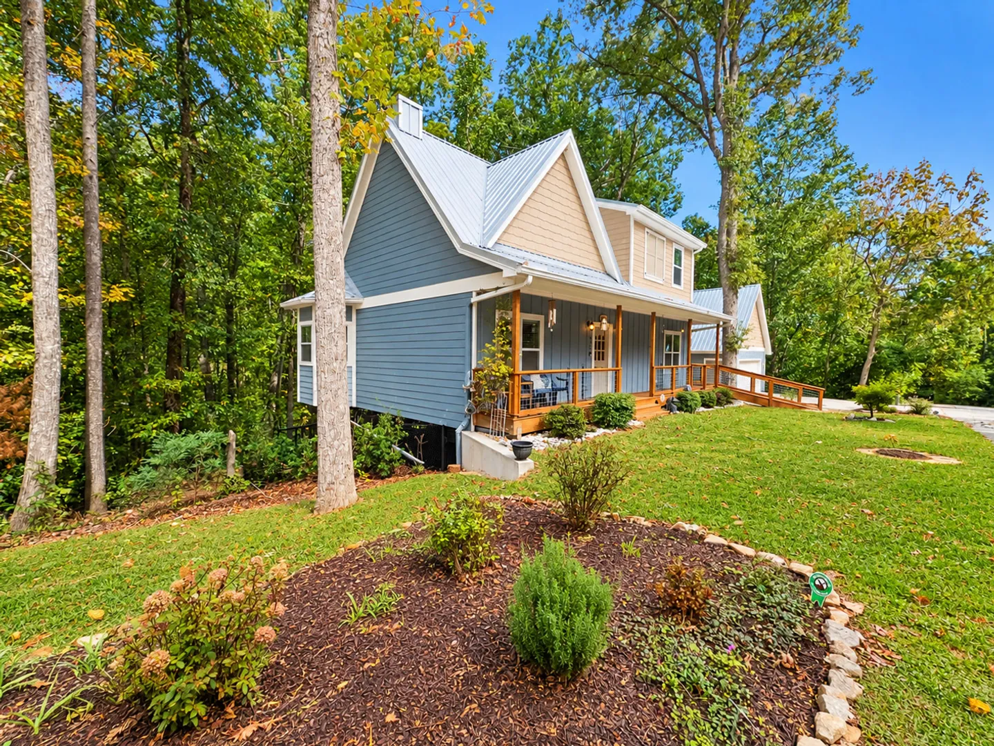 Exterior view of a blue and beige house with a metal roof, front porch, and wooded yard.