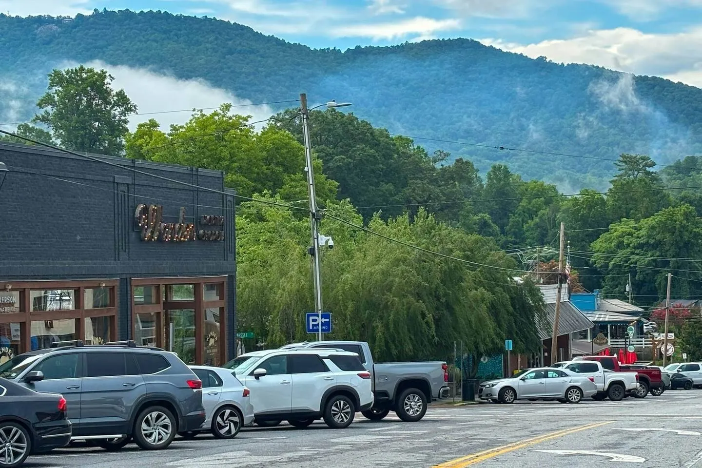 Street view of parked cars along a town road with brick storefronts and misty, forested mountains in the background.